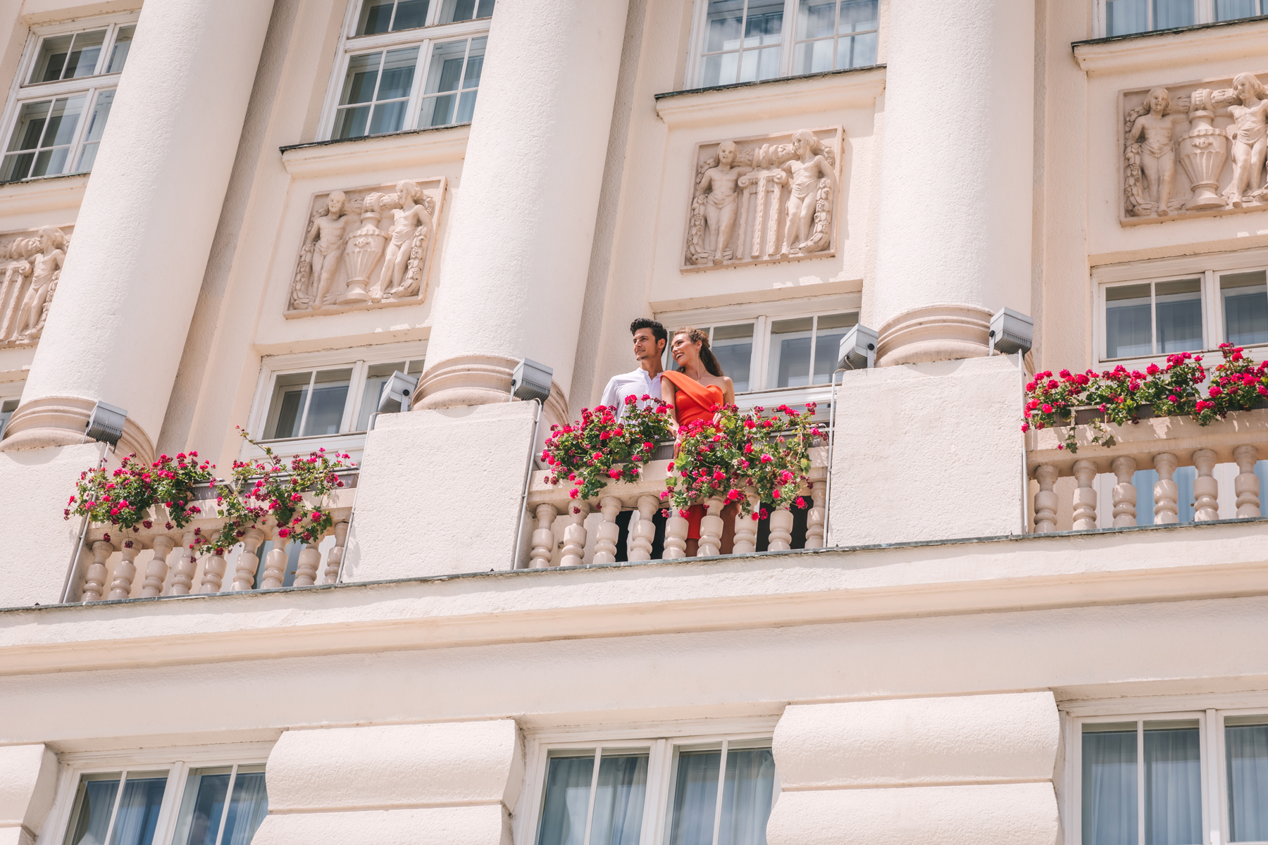 A couple standing on a balcony adorned with red flowers and classical architectural details.